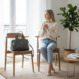 Woman sitting on a chair holding a cup in a bright room with a plant and handbag.