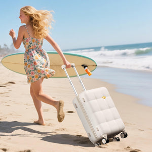 Woman with a floral dress and surfboard walking on a beach with a white suitcase.