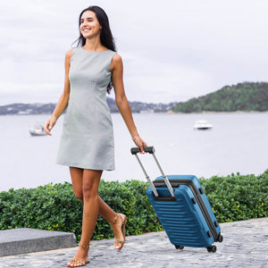 Woman in a light gray dress pulling a blue suitcase by a waterfront